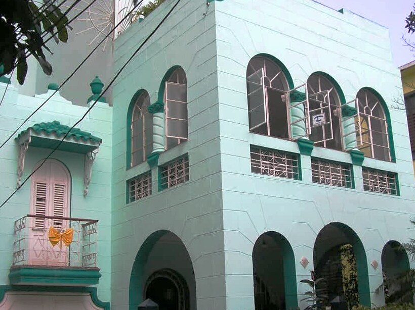 The exterior of a two-story building features a mint-green facade with decorative elements. Arched windows allow natural light to filter into the structure, and a small balcony is adorned with a yellow bow. Lush greenery surrounds the building, adding to its inviting appearance.