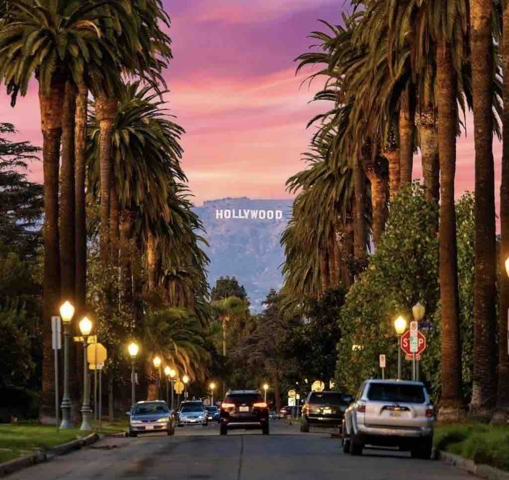 A tree-lined street is illuminated by street lamps, leading to the iconic Hollywood sign visible in the background against a pink and purple sky. Cars are parked along the road, adding a suburban feel to this picturesque setting.