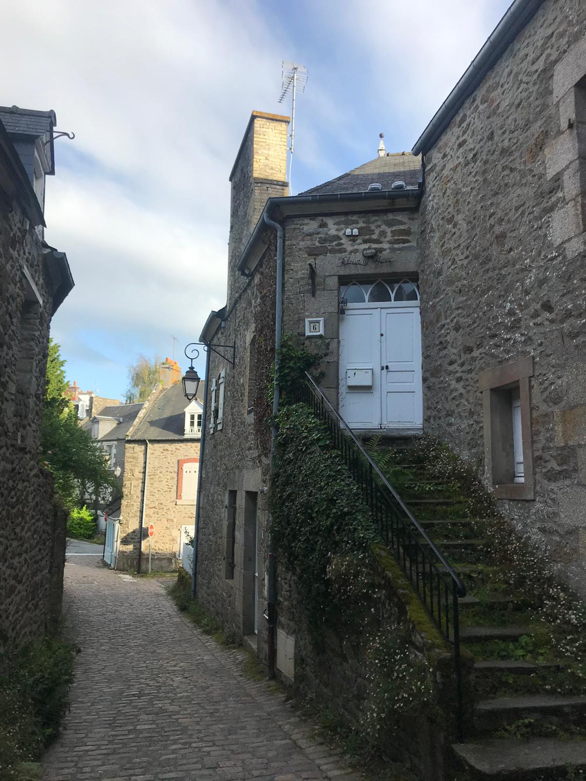 A narrow cobblestone path is shown leading toward a stone building. The entrance features a white door with decorative panels and is flanked by small windows. Green vines climb the side of the building, and a staircase leads up to the entrance. Soft sunlight filters through the clouds.