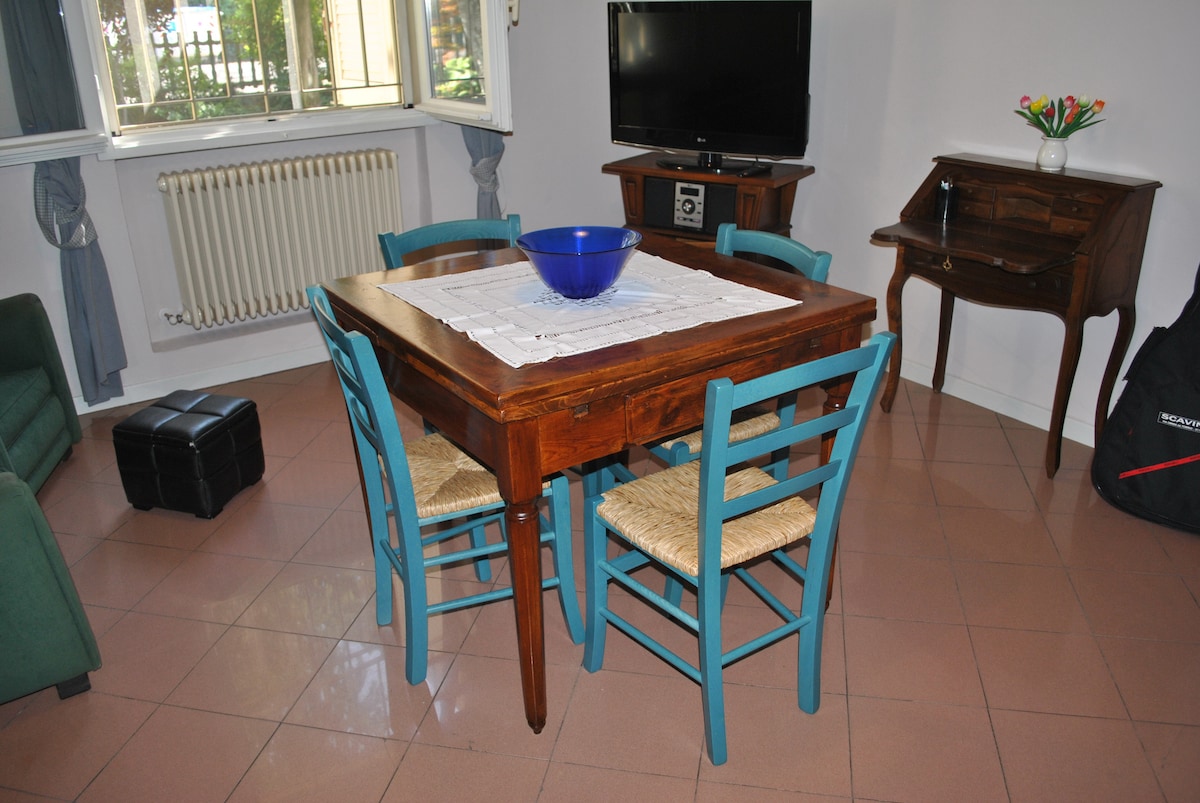 A wooden dining table is surrounded by three blue chairs with woven seats. A large blue bowl sits on the table, which is topped with a white table runner. A television and a small cabinet are visible in the background, along with partially draped windows allowing natural light.