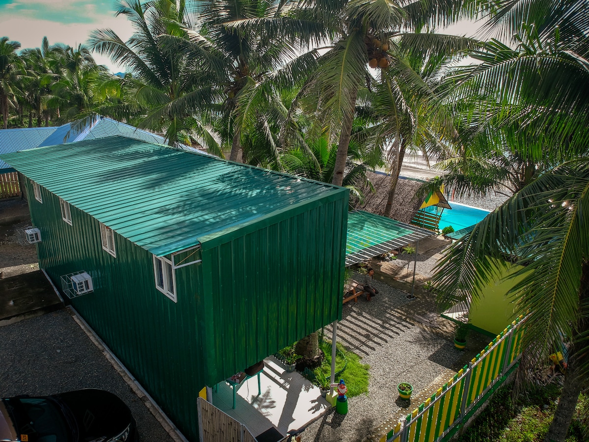 A green house with a corrugated metal roof is surrounded by tall palm trees. The outdoor space features shaded seating areas, with glimpses of an infinity pool and the beach visible in the background, creating an inviting atmosphere for relaxation.
