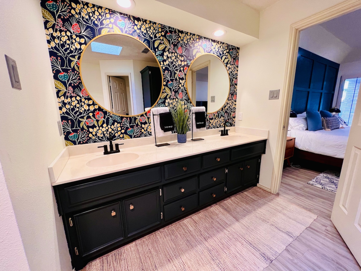 A spacious bathroom features dual sinks set against a floral-patterned accent wall. A stylish dark cabinetry provides ample storage beneath a long countertop. Natural light filters in from a skylight, enhancing the serene atmosphere. The floor is covered with a soft rug leading towards the bedroom.