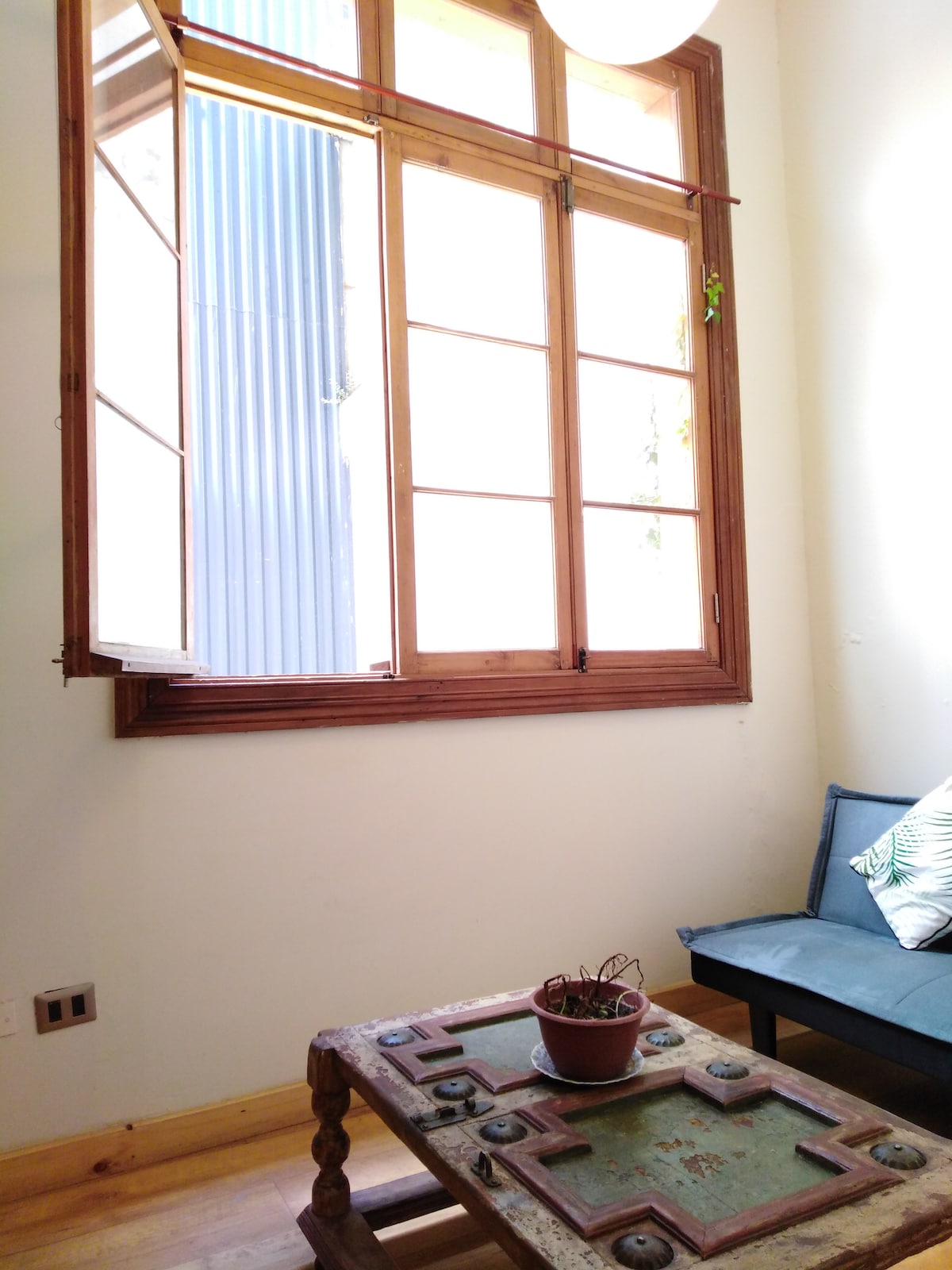 A window with wooden frames allows natural light to fill the corner of the room. A small, rustic table is positioned nearby, featuring decorative elements. A simple chair with a cushion complements the space, inviting relaxation near the sunlit window.