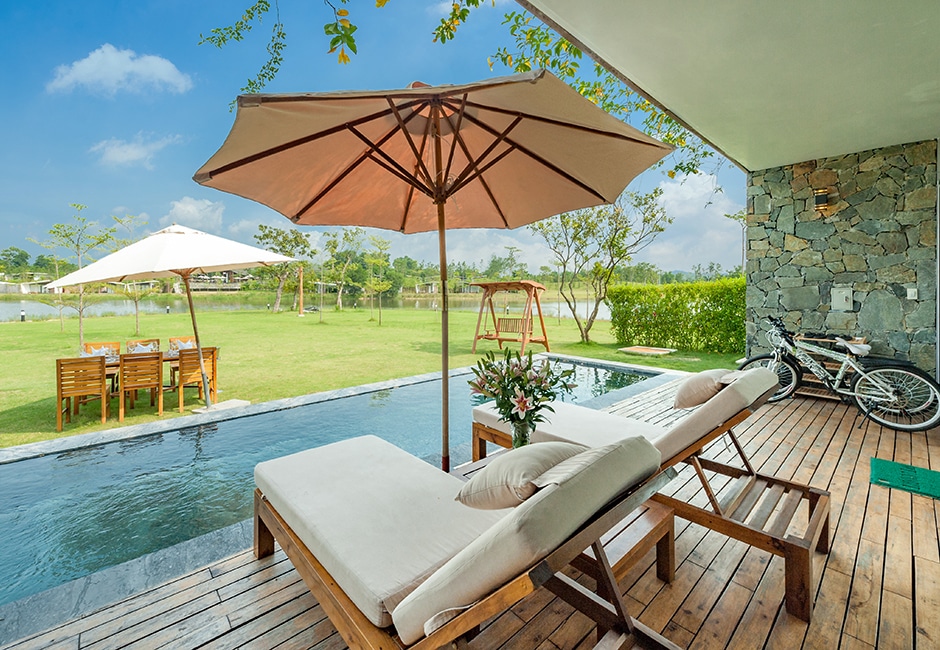 A tranquil outdoor area features two lounge chairs beside a narrow swimming pool, set against lush greenery. An umbrella provides shade, while a dining table with four chairs and a swing are visible in the background. A bicycle is positioned nearby on paved decking.