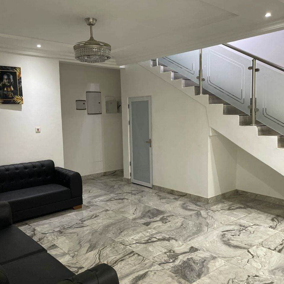 A modern living area is furnished with a dark sofa, while elegant marble flooring enhances the space. A staircase with a glass railing leads to the upper level. Ambient lighting is provided by a stylish chandelier, creating a bright and inviting atmosphere.