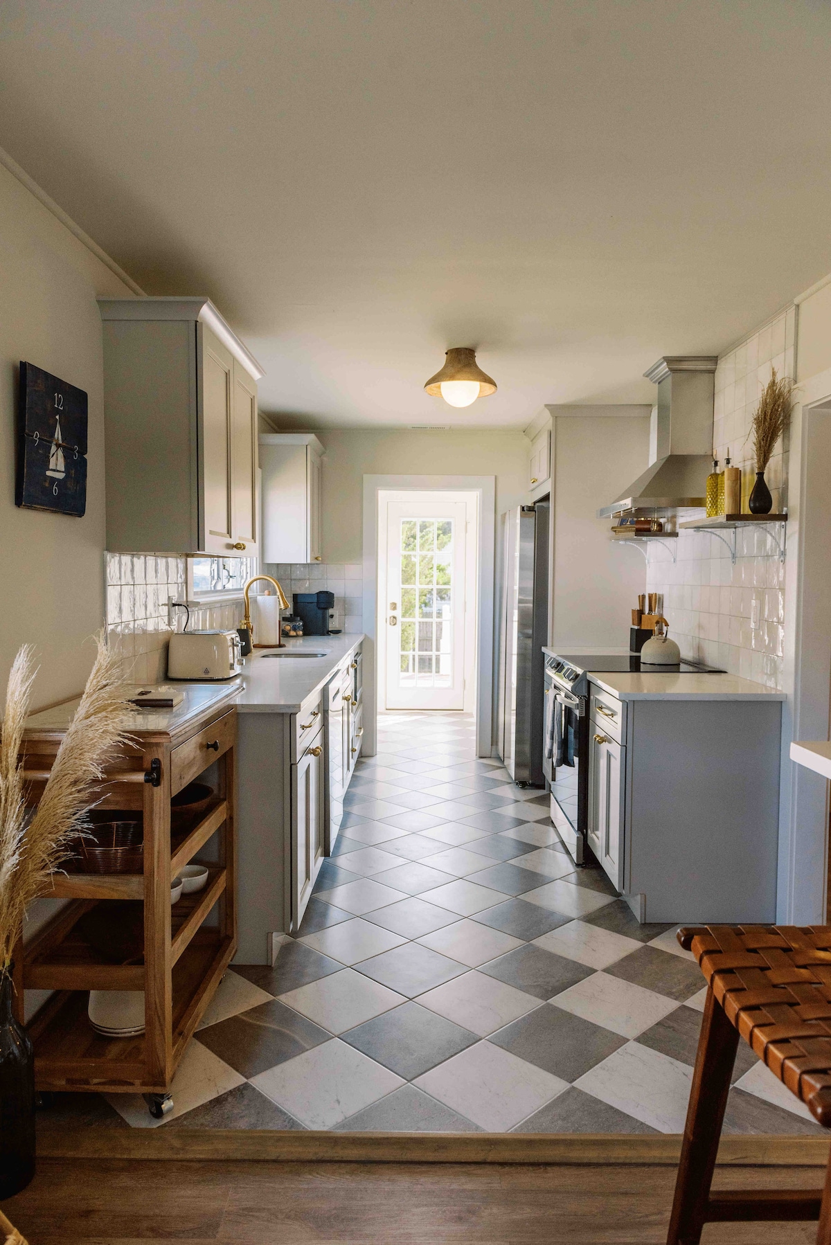 A modern kitchen features sleek cabinetry in soft grey tones, with a mixture of patterned and plain tiles underfoot. Large windows allow natural light to enter, illuminating the workspace. The area includes essential appliances and a small wooden cart, enhancing functionality and convenience.