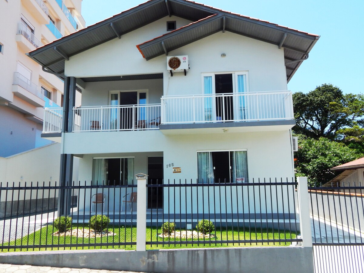 The exterior of a large, two-story house is displayed, featuring a light-colored facade and a sloped roof. A small garden is visible in front, along with a fenced area. Balconies are present on the upper floor, providing ample outdoor space.