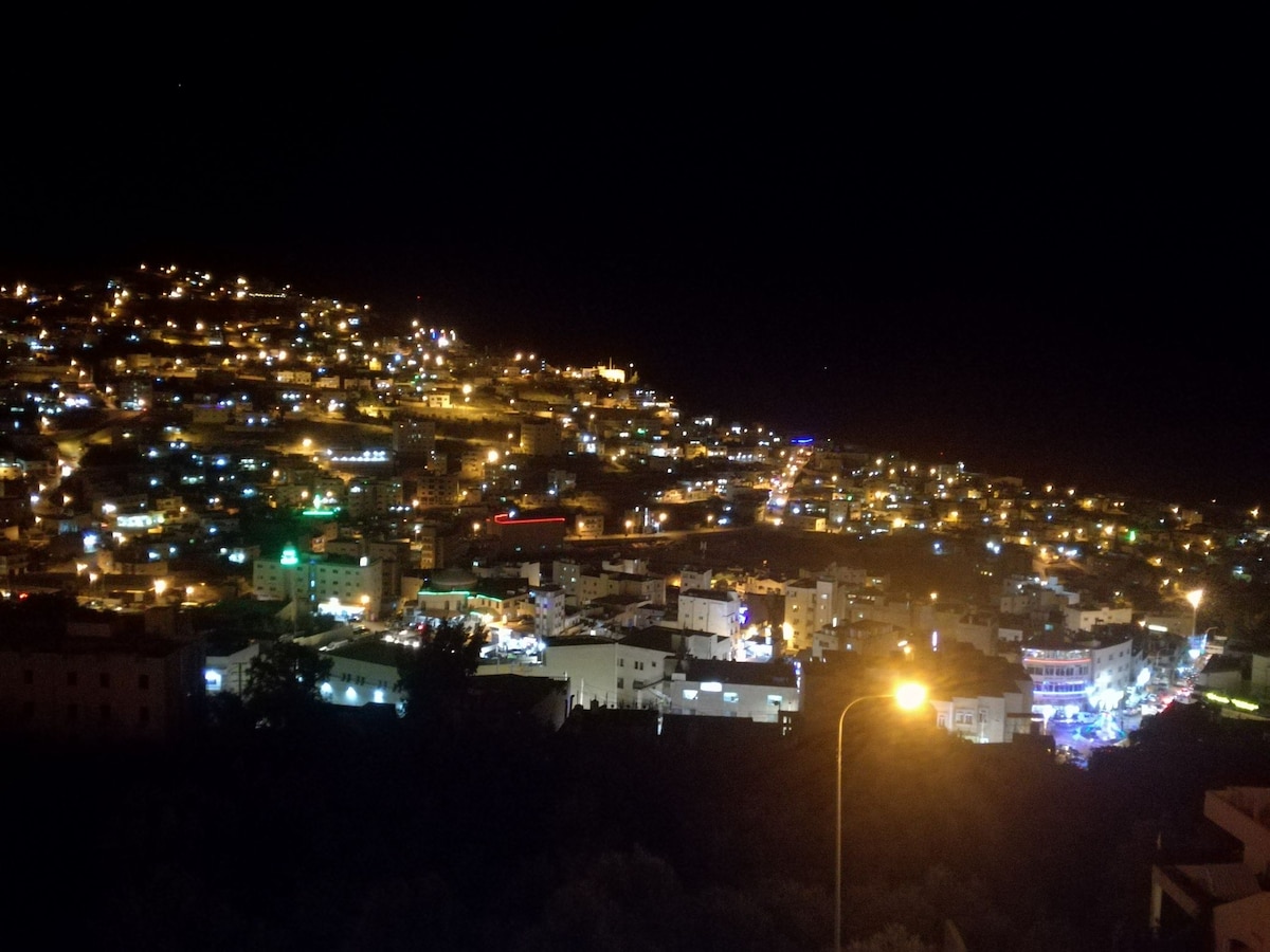 A nighttime view of a hillside town is illuminated by numerous lights, creating a vibrant scene. Buildings and streets are visible, with the glow of streetlights adding warmth. The landscape showcases layers of structures cascading down the slope, highlighting the town's unique layout.