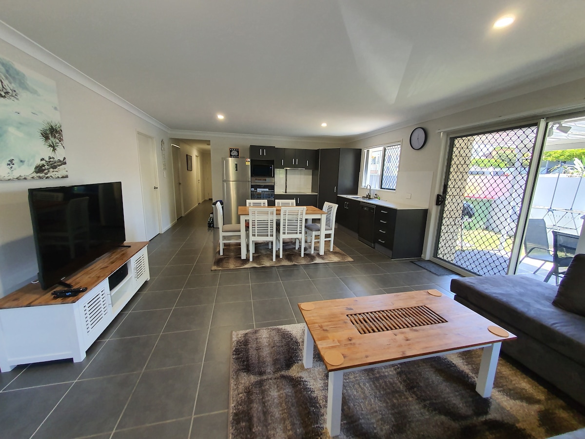 An open-plan living area features a large dining table surrounded by six chairs, with a modern kitchen visible in the background. A cozy sectional sofa sits adjacent to a wooden coffee table atop a textured rug. Large sliding doors provide access to an outdoor space.