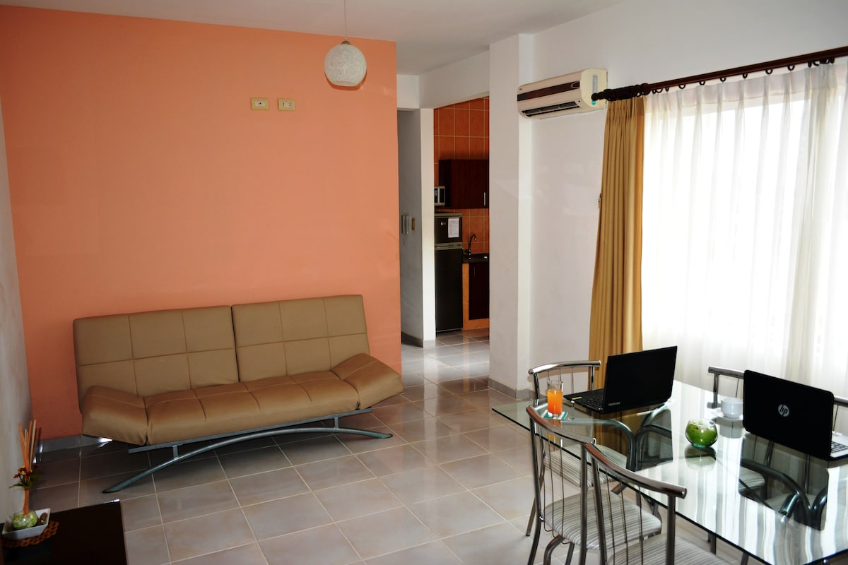 A living area features a light-colored sofa bed positioned against a peach wall. A glass dining table is accompanied by metal chairs, and a small bowl with decorative items adds a touch of detail. Natural light streams through sheer curtains, enhancing the space's brightness.