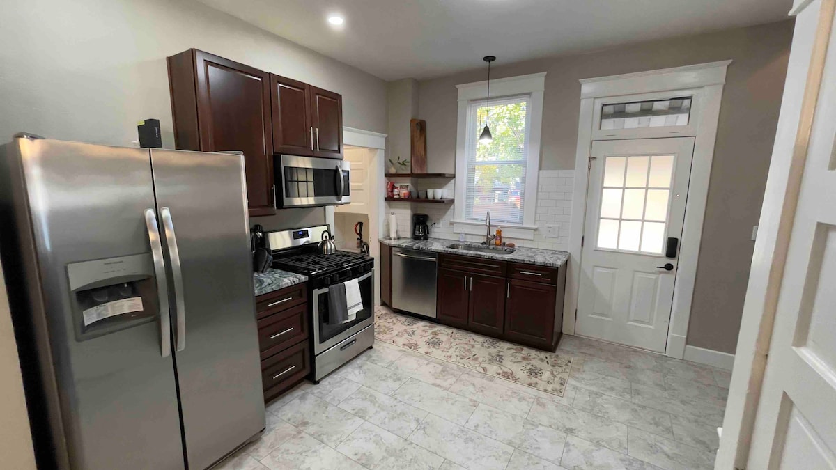 The kitchen features dark wood cabinets paired with granite countertops. Stainless steel appliances include a refrigerator and stove, while a door leads to the outside. Natural light fills the space through a nearby window, enhancing the neutral tones of the decor.