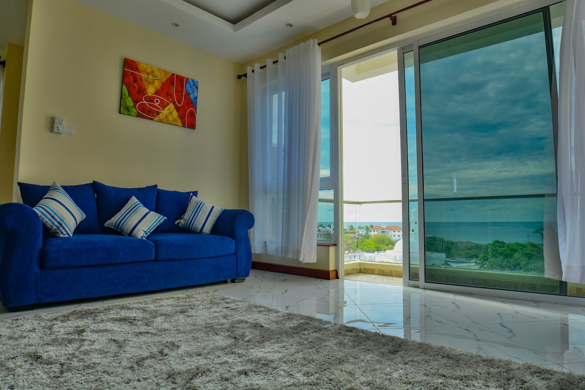 A bright living area features a blue sofa adorned with striped cushions. Large glass doors open to a balcony with expansive ocean views. Light-colored curtains frame the entrance, and a soft, plush rug lies on polished marble flooring, enhancing the room's airy atmosphere.