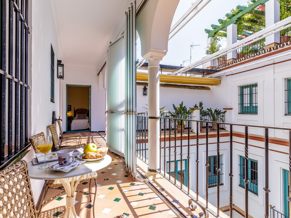 A private terrace features a small round table with two woven chairs, adorned with a breakfast setting. The terrace overlooks a beautifully designed inner patio with lush greenery and decorative tiles. Light filters through the partially open sliding glass doors, enhancing the serene ambiance.