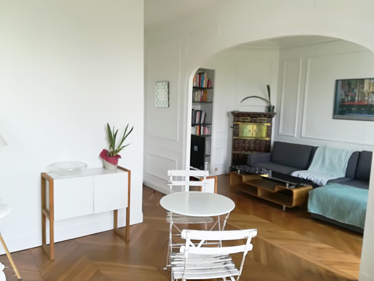 A bright living space is displayed, featuring a cozy seating area with a sofa and a light teal throw. A white round dining table is complemented by two metal chairs. A bookshelf is visible in the background, adding a touch of warmth through its wooden finish.
