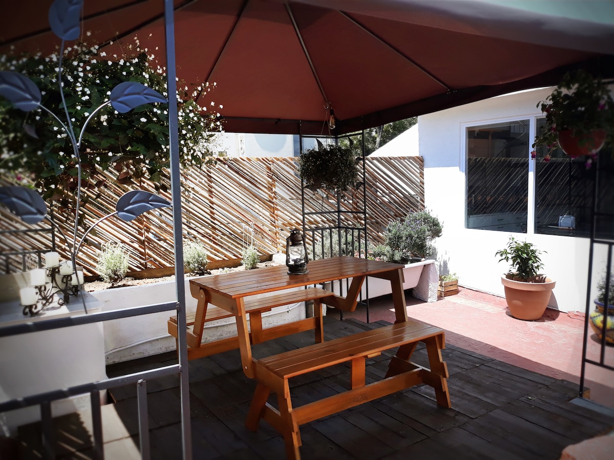 A shaded outdoor seating area features a wooden picnic table surrounded by planters with greenery. The space is enclosed by a wooden barrier and adorned with hanging plants, creating a serene environment for relaxation or dining.