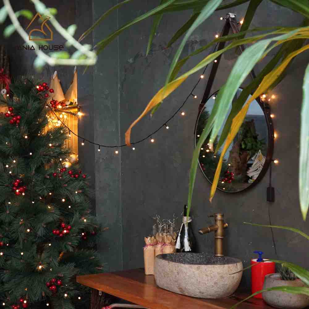A modern bathroom sink made of stone is positioned against a dark wall, reflecting warm light from string lights. A large round mirror enhances the space, and a festive green tree adorned with red decorations stands nearby, creating a cozy atmosphere.