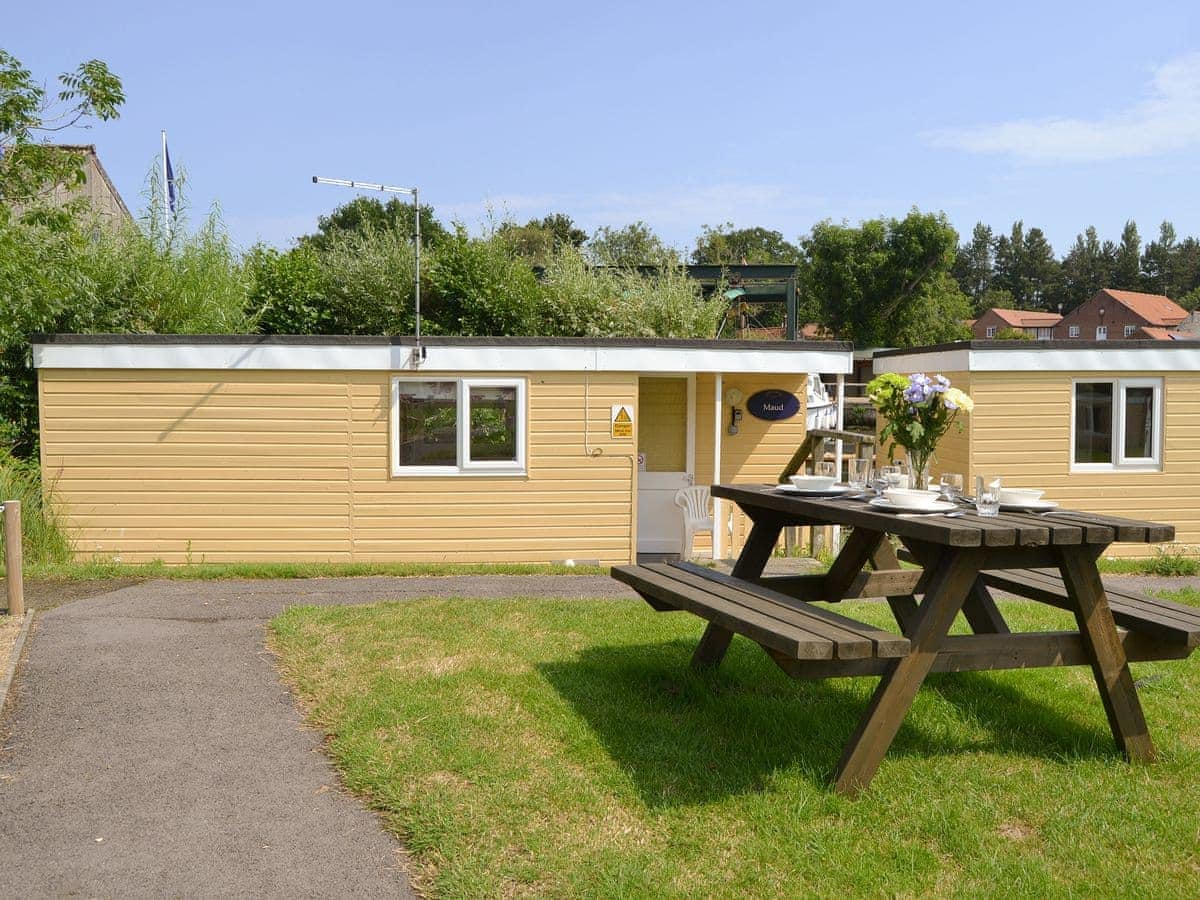A yellow houseboat is set amongst greenery, featuring a pathway leading to its entrance. A wooden picnic table is placed on the grass in the foreground, accompanied by tableware and a vase of flowers, inviting outdoor dining in a serene environment.