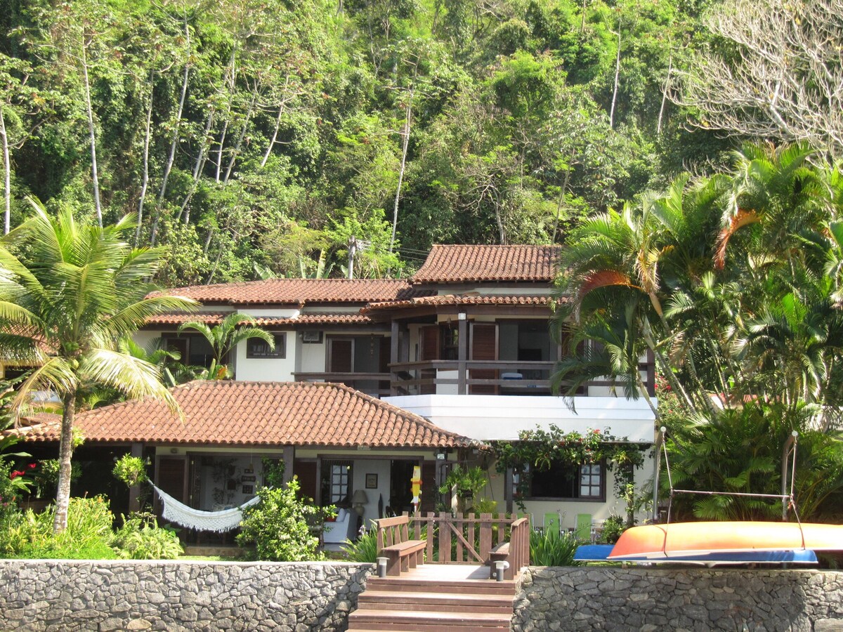 The oceanfront residence is set amidst lush greenery, with a two-story structure featuring traditional architecture and terracotta roof tiles. A wide wooden deck leads towards the entrance, surrounded by tropical plants and palm trees, creating a natural, inviting atmosphere.