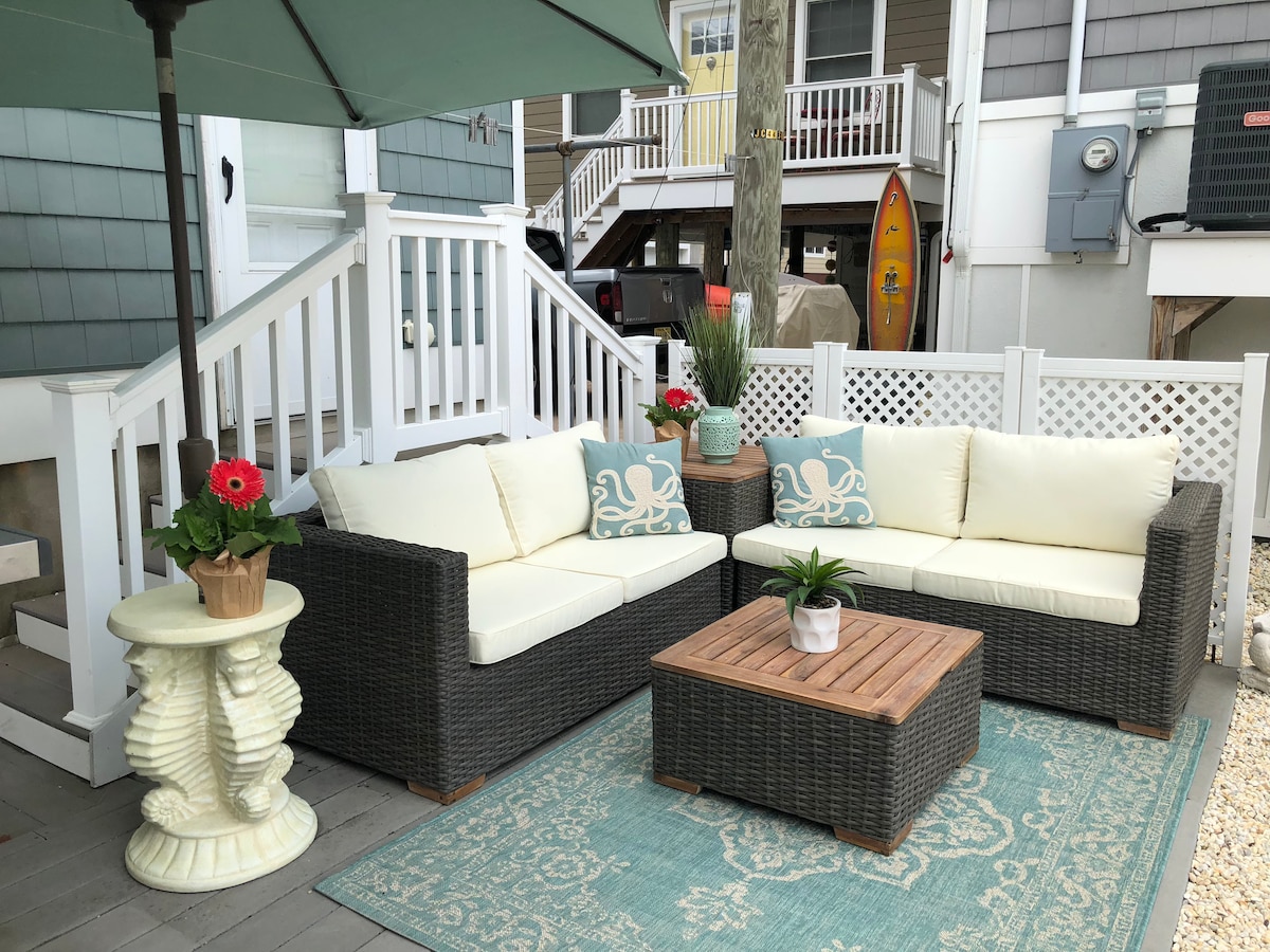 An outdoor seating area is showcased with a comfortable wicker couch adorned with cream cushions and decorative pillows. A wooden coffee table sits in the center, accompanied by potted plants and vibrant flowers. Surrounding elements include a patterned rug and an umbrella providing shade.