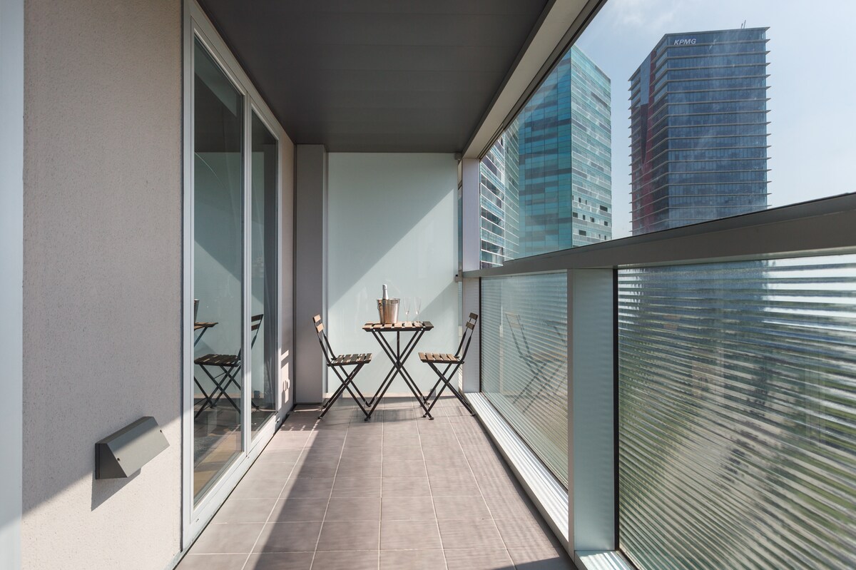 An outdoor balcony features a small round table set with two folding chairs. Floor-to-ceiling glass panels offer unobstructed views of surrounding modern buildings. Neutral-toned tiles cover the floor, and sunlight filters through, creating an inviting space for relaxation.