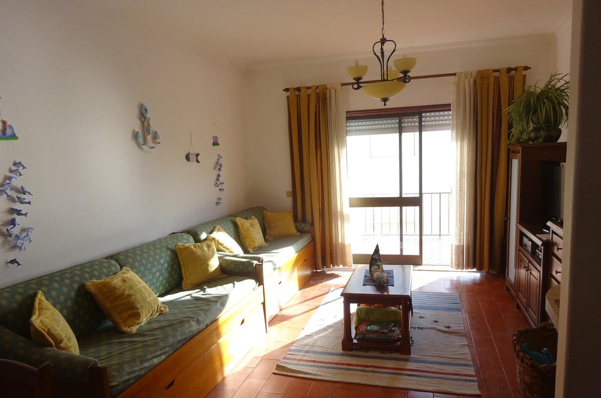 A spacious living area features two comfortable sofas adorned with yellow cushions. Natural light enters through a glass sliding door leading to a balcony. A wooden coffee table sits in the center, complemented by a colorful rug and decorative wall elements.