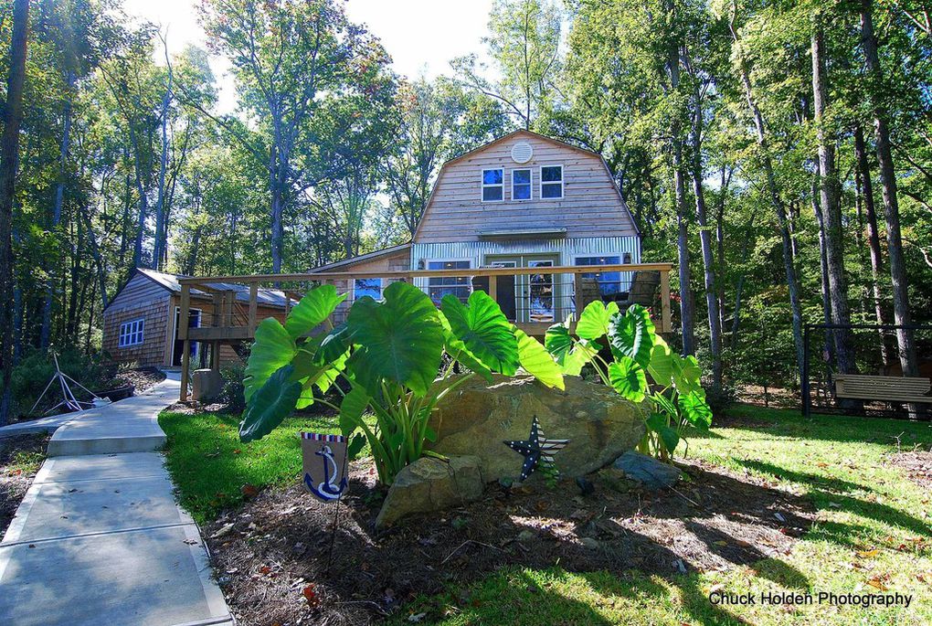 A rustic cabin is surrounded by lush greenery, with a stone pathway leading to a large deck. Large, tropical-looking plants frame the entrance, while the cabin features multiple windows. The structure presents a charming blend of wood siding and natural elements.