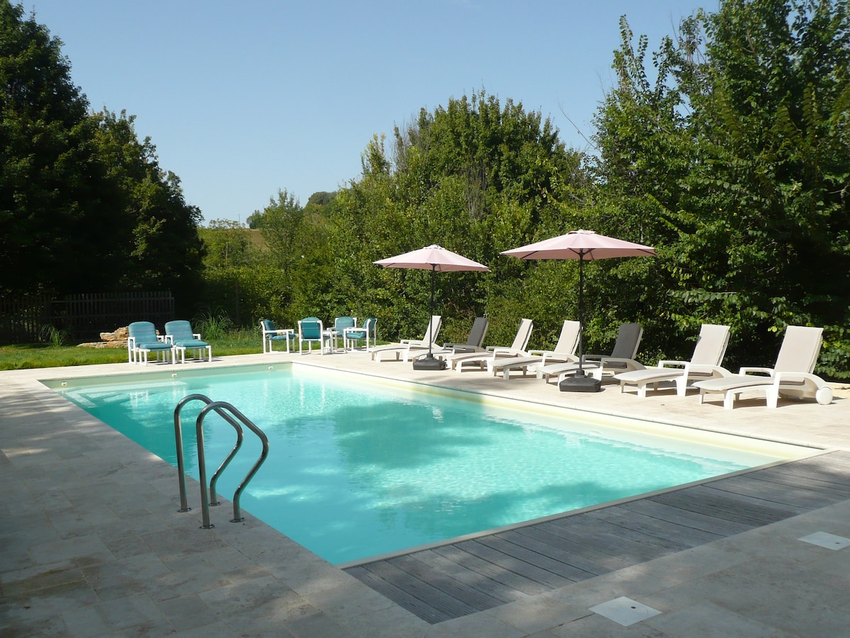 The outdoor pool is surrounded by landscaped gardens, featuring a large swimming area with a gentle gradient. Comfortable loungers and umbrellas are positioned around the pool, providing shaded spots for relaxation. The tranquil setting is enhanced by greenery visible in the background.