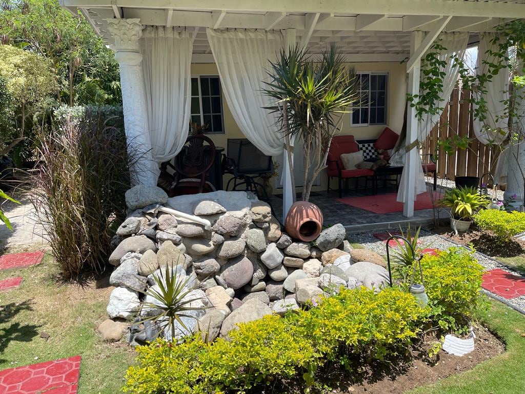 A landscaped garden area features a rock formation with a decorative pot, surrounded by carefully arranged greenery and plants. A shaded seating area with cushions is visible, alongside flowing curtains draping from the roof, creating an inviting space for relaxation.
