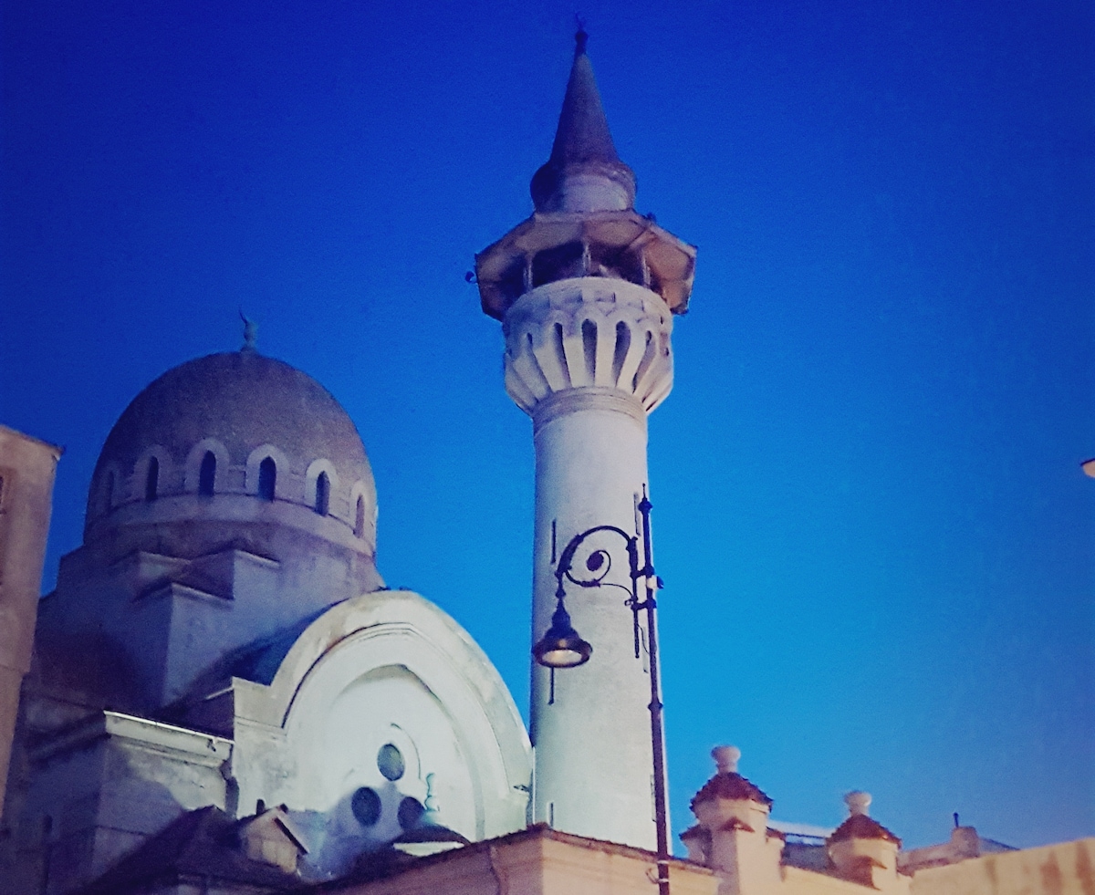 A historic building is featured, showcasing a prominent minaret topped with a pointed roof and a dome. The intricate architectural details are highlighted against a twilight sky, enhancing the structure's elegance. Street lighting is visible, adding to the serene evening ambiance.