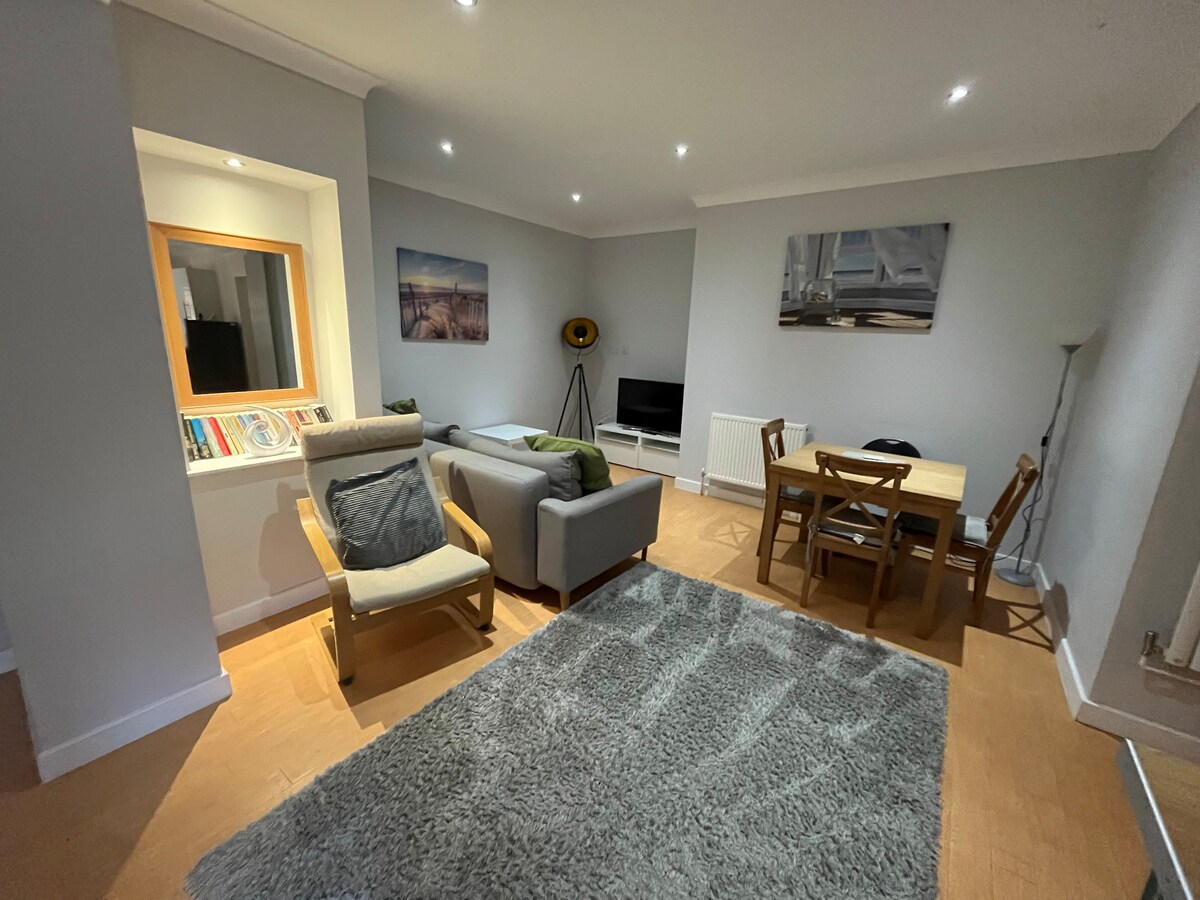 An open plan lounge area features a corner sofa, an armchair, and a dining table with four chairs. Natural light brightens the space, complemented by soft gray walls. A television is positioned against a wall, while decorative artwork adds a subtle touch.