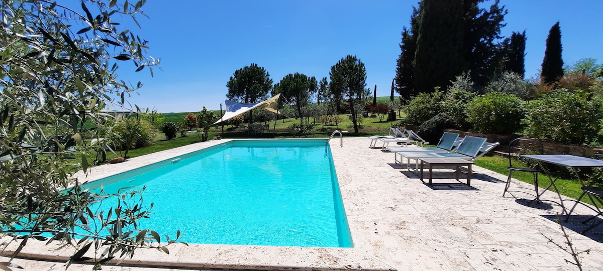 The private swimming pool glistens under clear blue skies, surrounded by a beautifully landscaped garden. Sun loungers and seated table areas are arranged around the pool, providing ample space for relaxation and outdoor dining.