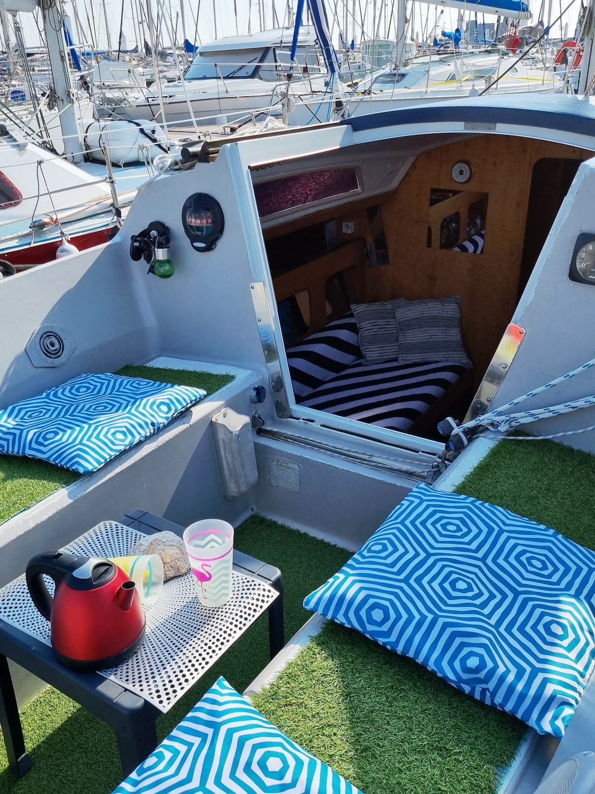 An outdoor seating area is visible on the boat, featuring two green mats and patterned cushions. A small table carries a kettle and two cups, with decorative stones nearby. The entrance to the cabin is seen in the background, leading into a cozy, wooden interior.