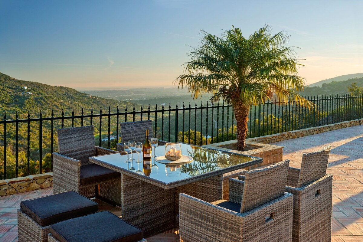 An outdoor dining area is displayed with a glass-topped table surrounded by wicker chairs. A palm tree stands nearby, and distant hills can be seen against the backdrop of a sunset sky. The scene conveys a relaxed ambiance for outdoor dining.
