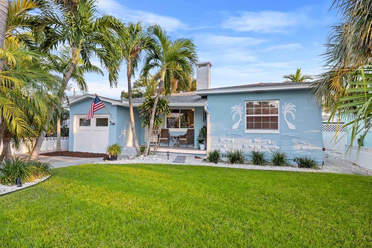 A charming beach house is depicted, featuring a light blue exterior with palm tree motifs. Lush green grass surrounds the property, with well-maintained landscaping and palm trees lining the entrance. An American flag is displayed on the porch, along with seating for relaxation.