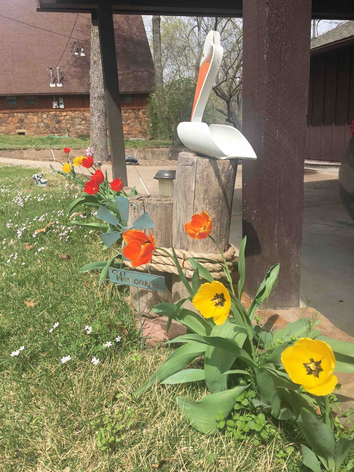 A decorative wooden welcome sign is positioned near colorful flowers, including yellow tulips and red blossoms, at the entrance of the cabin. A white pelican sculpture sits on a wooden post, adding a whimsical touch to the welcoming ambiance of the outdoor space.