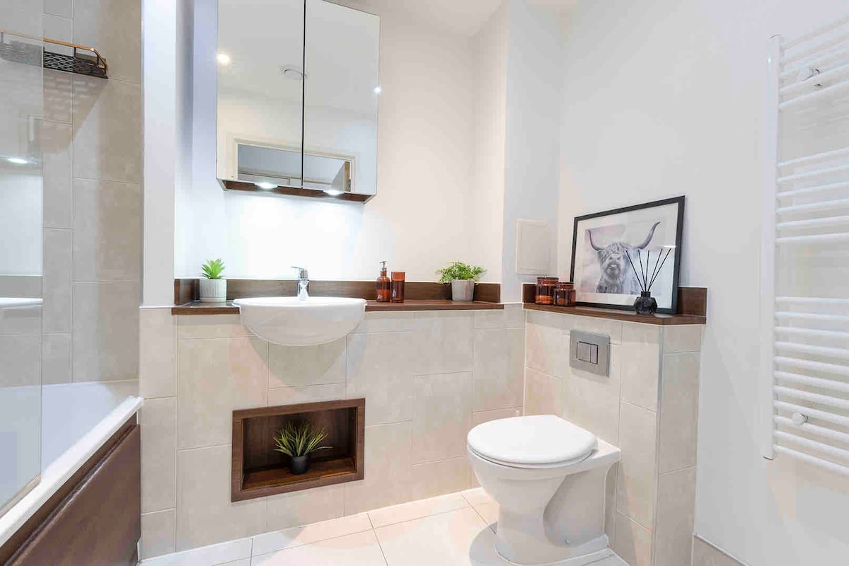 A modern bathroom features a wall-mounted sink above a wooden shelf. A sleek mirror reflects soft lighting. Decorative plants and candle holders accentuate the space. A towel radiator is positioned to the right of the toilet, enhancing the contemporary design.