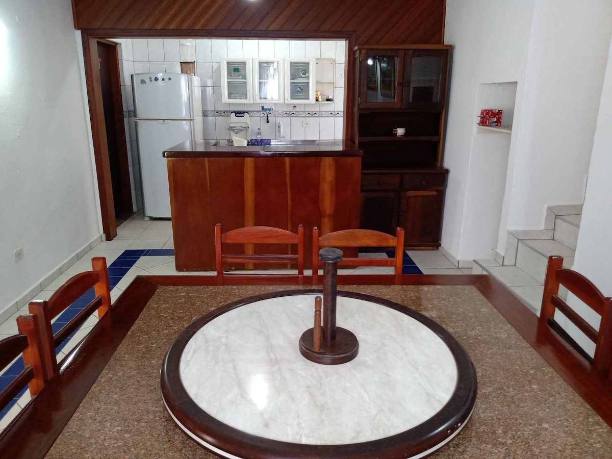 A dining area features a round marble table with a wooden centerpiece and six wooden chairs. In the background, a kitchen is visible with white cabinetry and appliances, while a bar counter adds an additional eating space.