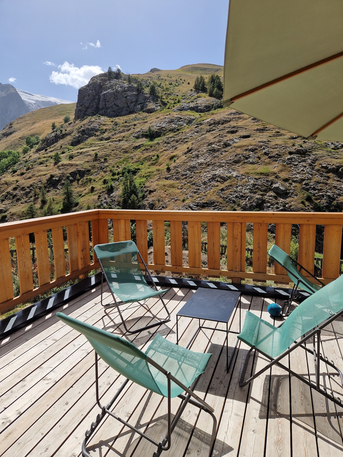 A terrace features wooden decking and a protective wooden railing, overlooking scenic mountainous landscape. Two green chairs and a small table create a relaxing outdoor space, with a sunshade providing additional comfort. The abundant natural light highlights the surroundings.