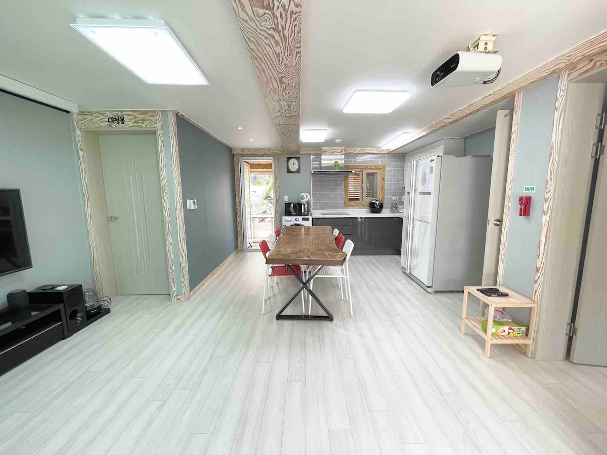 The spacious living area features a large wooden dining table surrounded by multiple chairs. A sleek kitchen equipped with modern appliances is visible in the background. Natural light fills the space through the nearby windows, enhancing the inviting atmosphere.