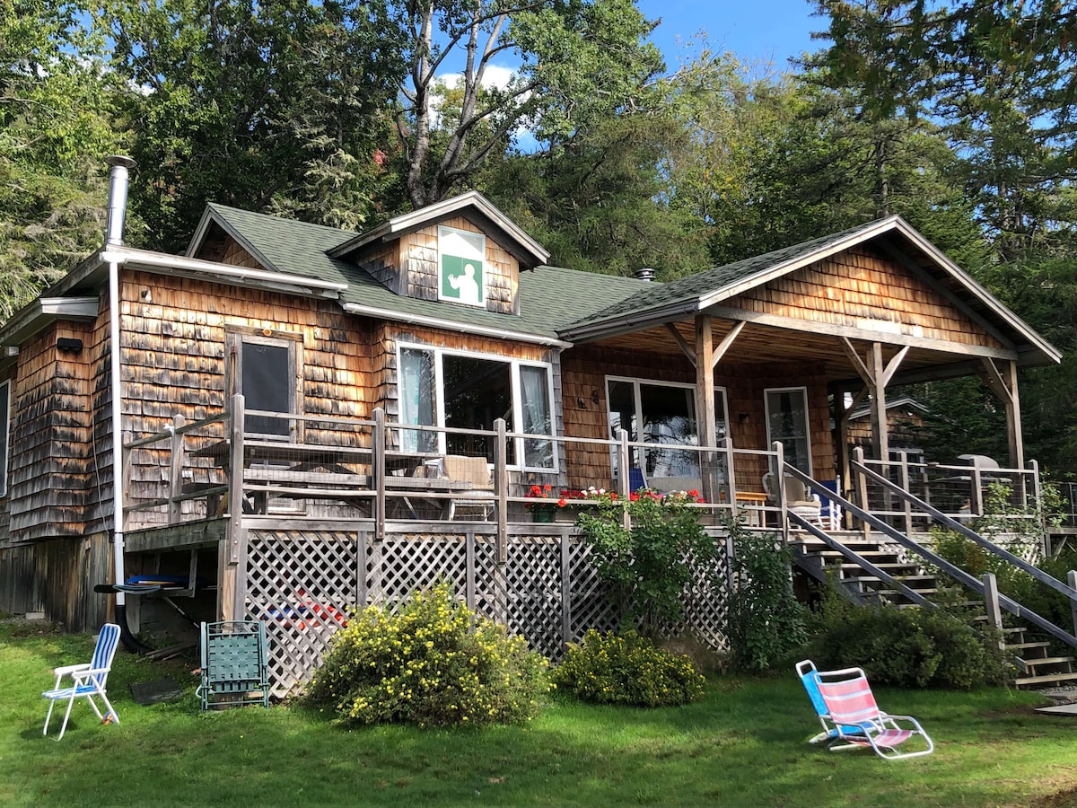 A rustic cabin exterior is showcased, featuring wooden siding and a wraparound porch. The entrance is adorned with a green cross sign. Surrounding the cabin are well-maintained lawns and greenery, along with a few colorful lawn chairs positioned in the grass.