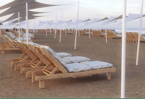 A sandy beach area is visible, featuring several wooden lounge chairs with cushions arranged in neat rows. Large white canopies provide shade over the seating area, enhancing comfort by the shoreline.