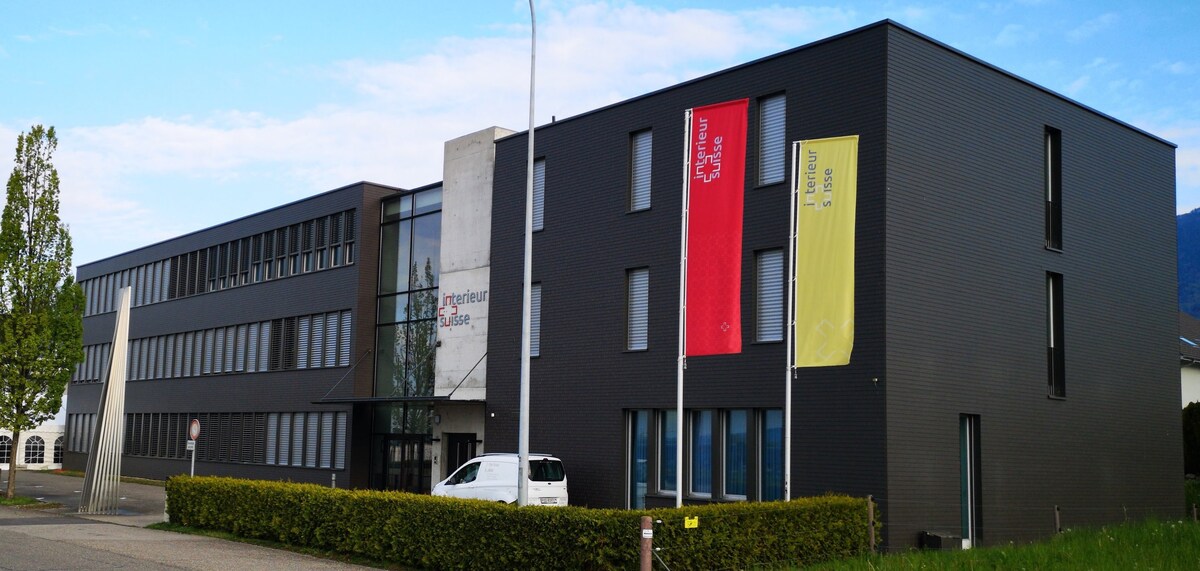 The modern building features a sleek black exterior with large windows and a minimalist design. Prominent flags in bright colors are displayed at the entrance, and a grassy area with neatly trimmed hedges surrounds the property. A vehicle is parked nearby.
