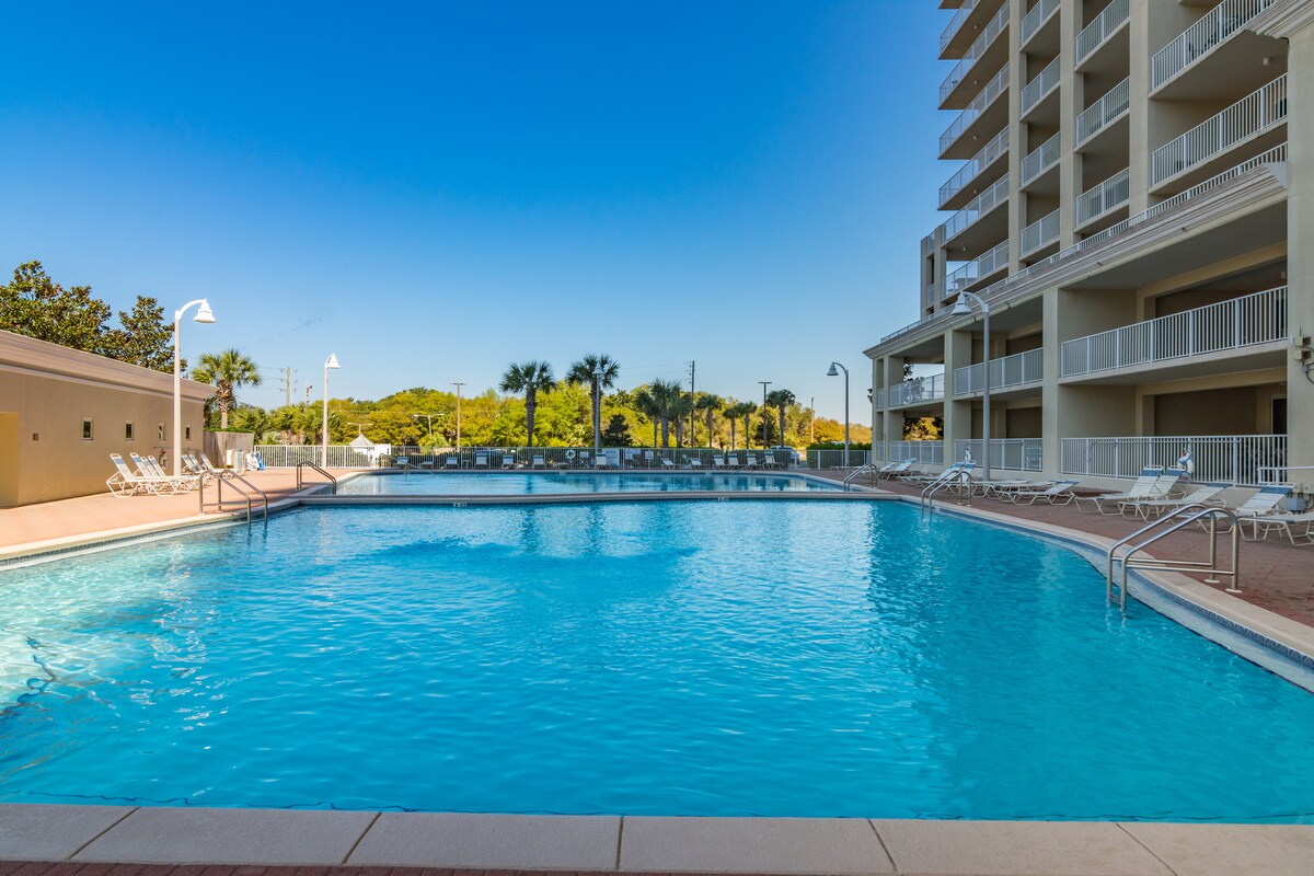 A large pool is surrounded by palm trees and lounge chairs, inviting relaxation under a clear blue sky. The pool's water sparkles in the sunlight, while a spacious building is visible in the background, enhancing the resort's inviting atmosphere.