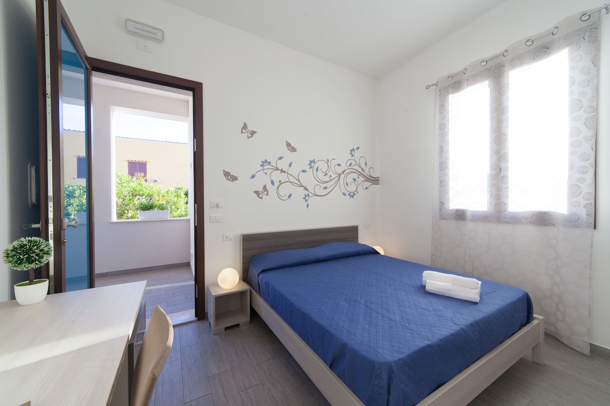 A simple and bright bedroom is presented with a spacious bed dressed in blue linens. Natural light enters through two windows, complemented by soft curtains. A small desk and a decorative plant add to the room's functionality, while a gentle wall design of flowers and butterflies enhances its charm.