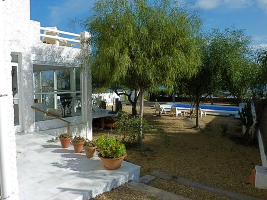 Lateral con entrada a la casa a la izquierda, jardín, piscina y el mar al fondo.