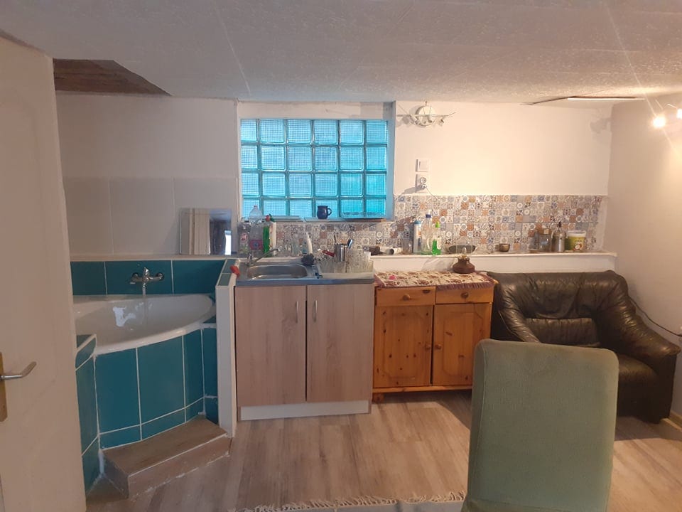 A compact kitchen area is displayed, featuring a small sink and a wooden cabinet with a countertop. Light blue tile contrasts with the patterned backsplash. A comfortable leather chair is positioned nearby, while a partially visible bath is situated on the left side of the image.