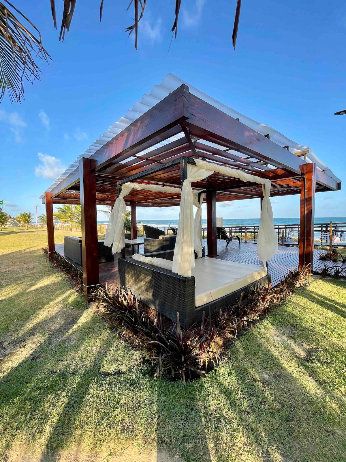 A shaded outdoor pavilion made of wood features open sides and a spacious seating area. Neutral-toned cushions are arranged on the seating, while lush grass surrounds the structure. Sunlight casts elongated shadows across the deck, creating a serene atmosphere near the water.