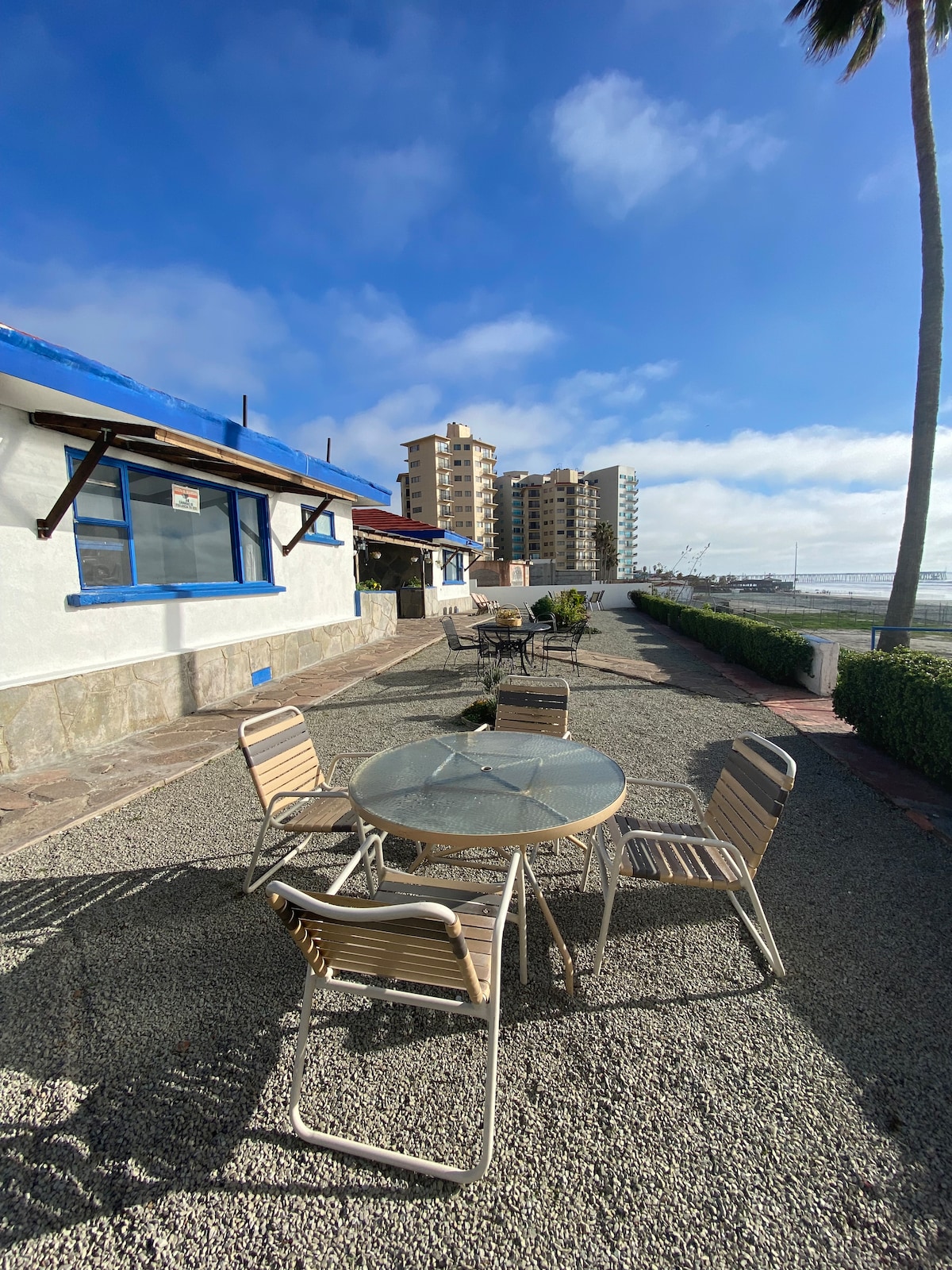 An outdoor seating area features a round glass-top table surrounded by four metal chairs, situated on a gravel pathway. Lush greenery lines the space, with tall palm trees providing shade, and a distant view of a beach and buildings under a clear blue sky.