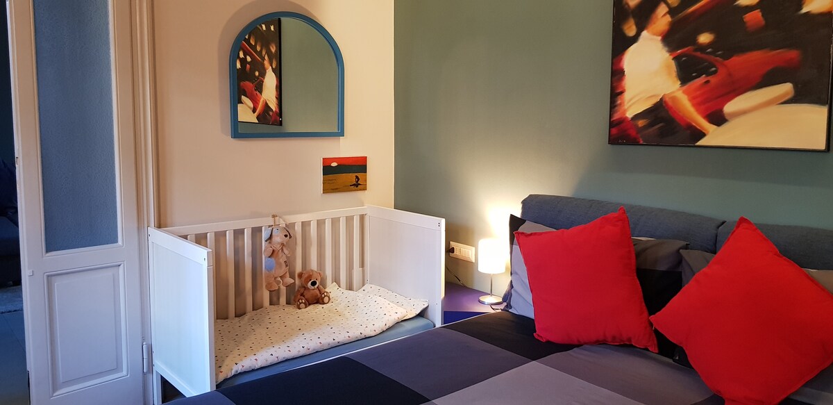 A cozy bedroom features a spacious bed adorned with colorful pillows. A small crib is positioned beside the bed, enhancing family-friendly accommodations. Soft lighting from a bedside lamp creates a calm atmosphere, while an abstract artwork adds a touch of modern design to the wall.
