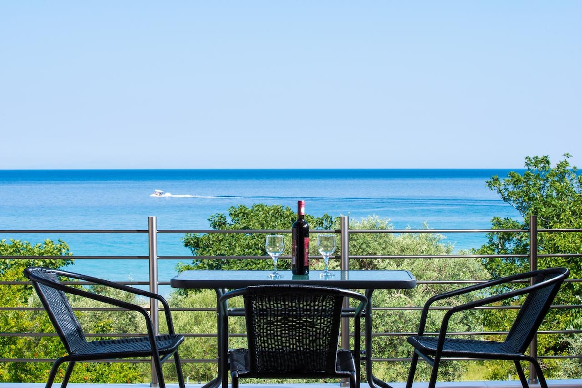 A balcony view showcases a table set with wine glasses and a bottle, overlooking a serene sea. Two black chairs frame the table, with vibrant greenery in the foreground and a clear blue sky enhancing the tranquil setting.